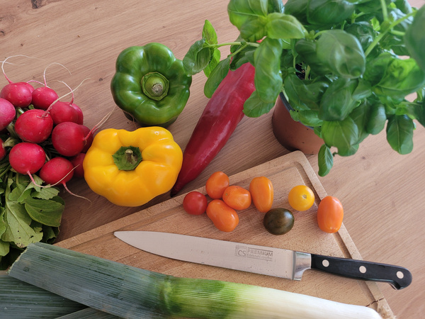 KI generiert: Frisches Gemüse, ein Messer und Basilikum auf einem Holzbrett. Radieschen, Paprika, Tomaten, Lauch.
