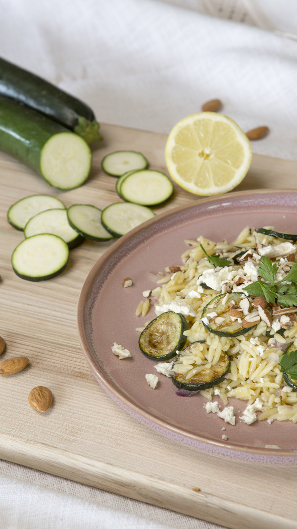 KI generiert: Ein Gericht mit Zucchini, Nudeln und Mandeln auf einem Teller, daneben eine halbe Zitrone und geschnittene Zucchini.