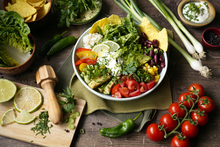 KI generiert: Das Bild zeigt eine bunte, mexikanisch inspirierte Bowl mit Zutaten wie Tomaten, Bohnen, Mais, Avocado und Tortilla-Chips. Drumherum sind frische Zutaten wie Limetten, Kirschtomaten und Frühlingszwiebeln verteilt.