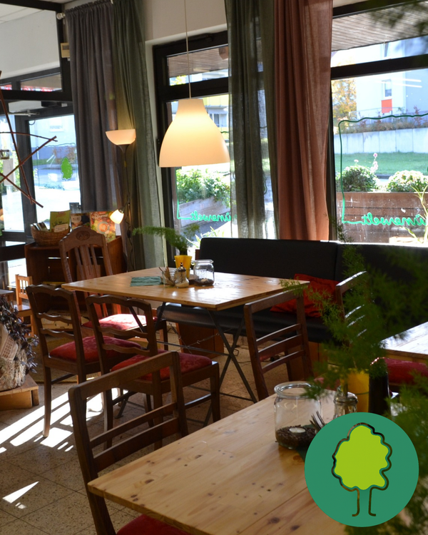 KI generiert: Gemütliches Café-Interieur mit Holzstühlen und Tischen, dekoriert mit Pflanzen. Ein Baum-Symbol ist dargestellt.
