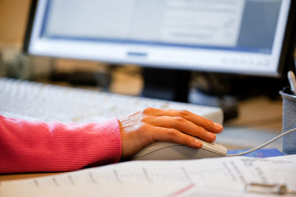 KI generiert: Eine Hand auf einer Computermaus mit unscharfem Bildschirm im Hintergrund. Vordergrund zeigt Papierdokumente.