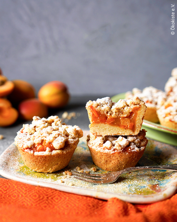 KI generiert: Aprikosen-Streuselmuffins auf einem Teller mit Gabel, Aprikosen im Hintergrund. Text: "Ökokiste e.V."