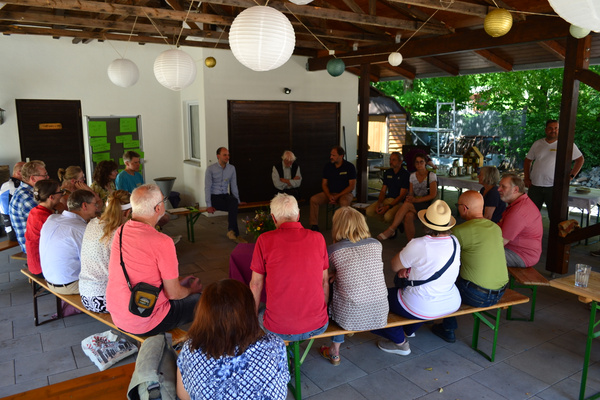KI generiert: Eine Gruppe von Menschen sitzt in einem Halbkreis unter einer überdachten Terrasse.