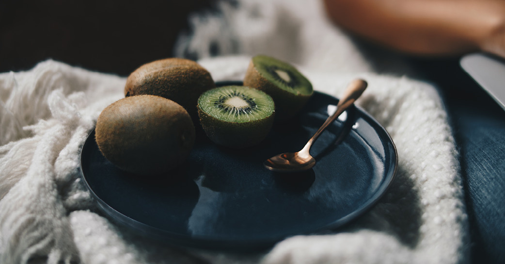 KI generiert: Kiwis und ein Löffel auf einem dunklen Teller, arrangiert auf einem weißen Stoff.