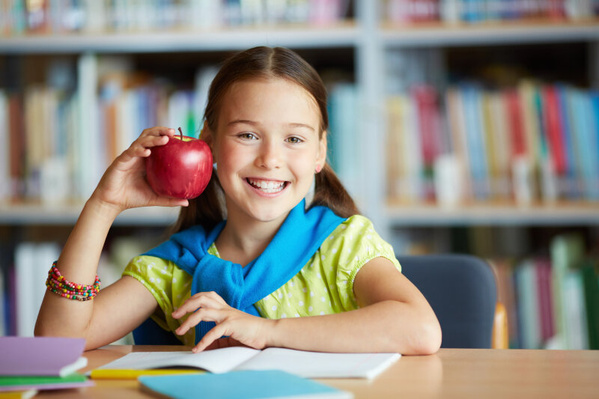 KI generiert: Mädchen in einer Bibliothek, hält einen roten Apfel und lächelt. Im Hintergrund sind Bücherregale zu sehen.